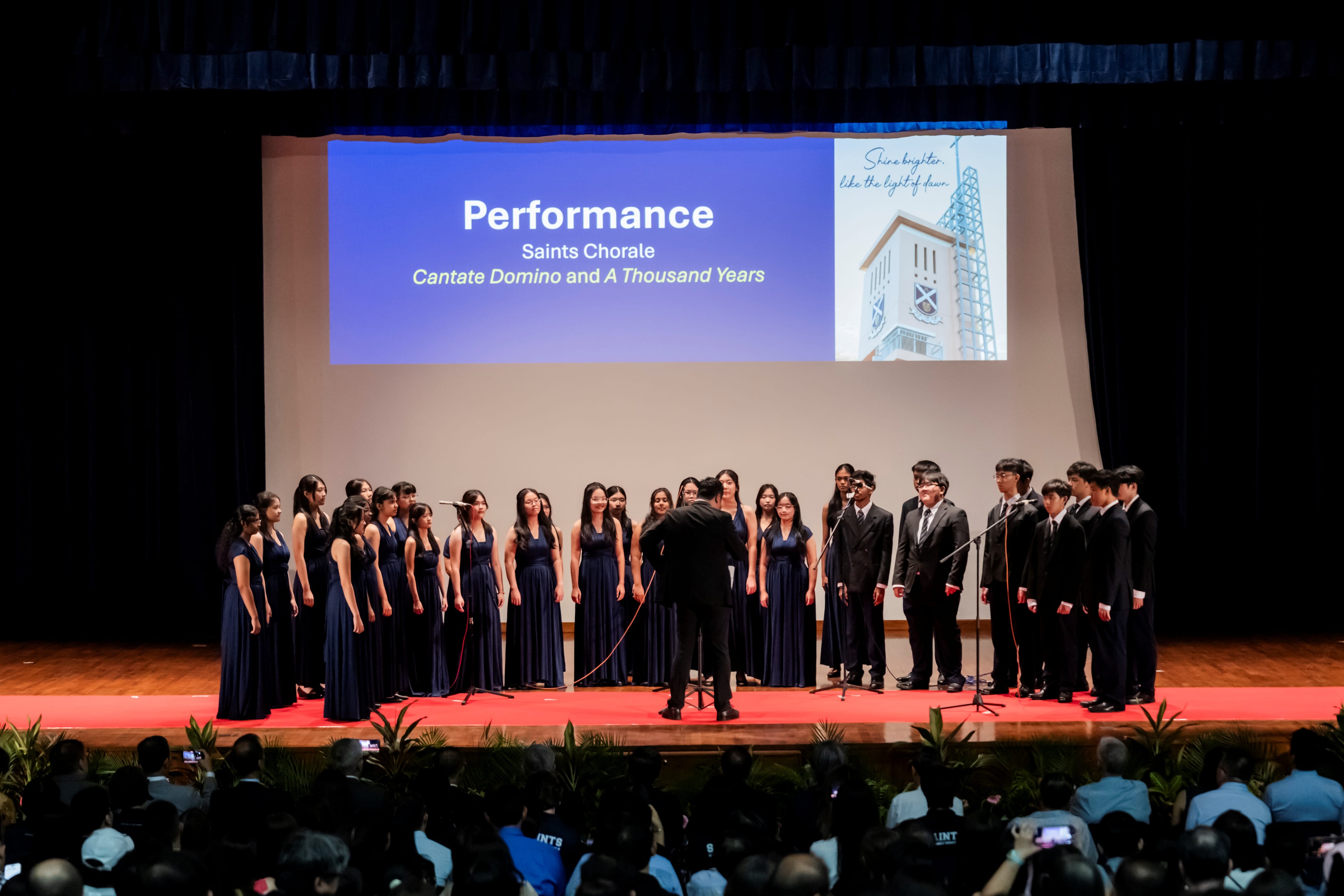 The Saints Chorale choir performs on stage under the direction of a conductor, beneath a projection screen displaying their performance piece titles.